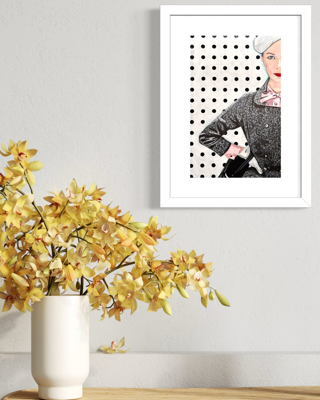 Framed limited edition print of a woman with a polka dot background next to a vase with yellow flowers on a wooden surface.
