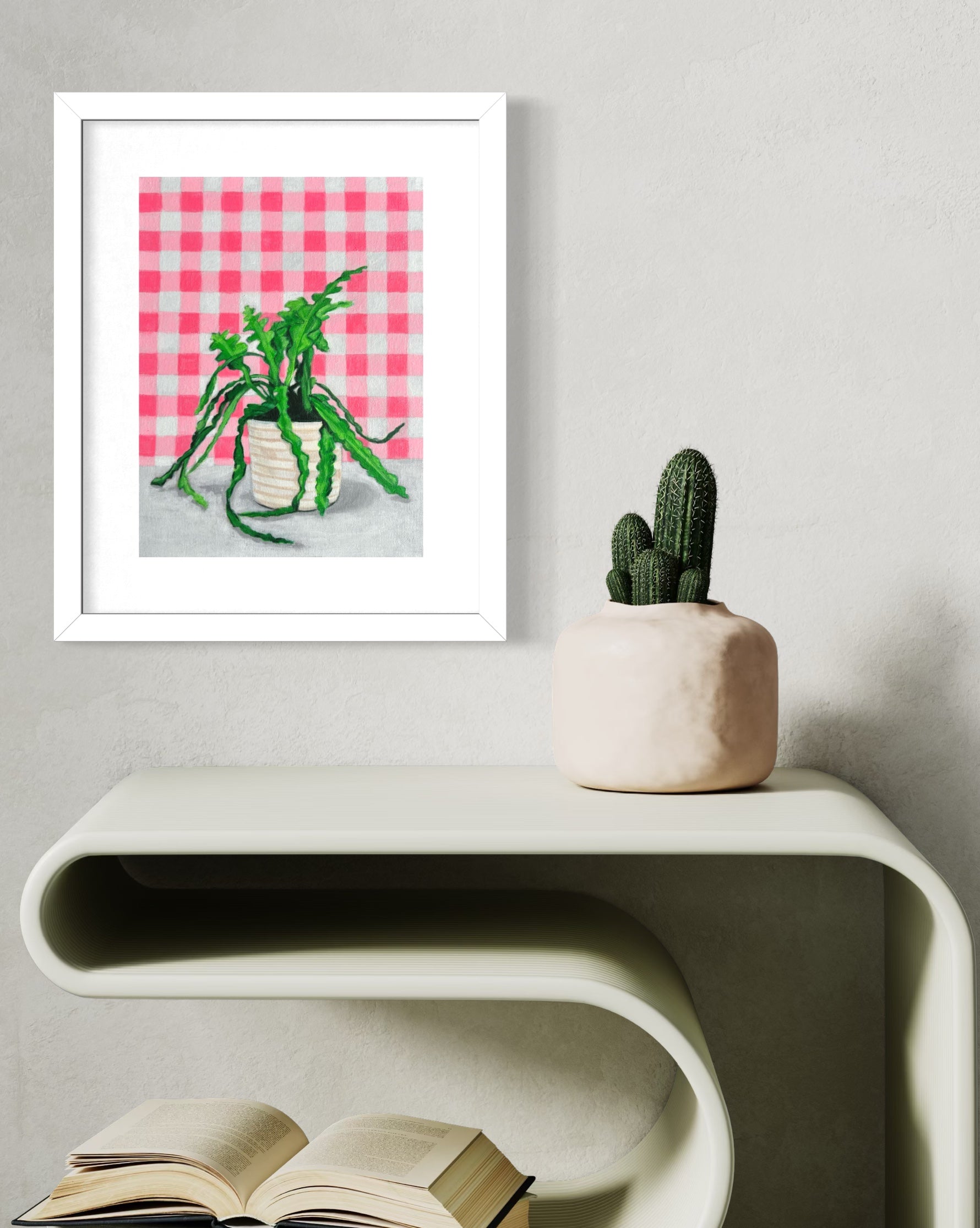 Framed limited edition print of a plant on a wall above a table with a book and a potted plant.