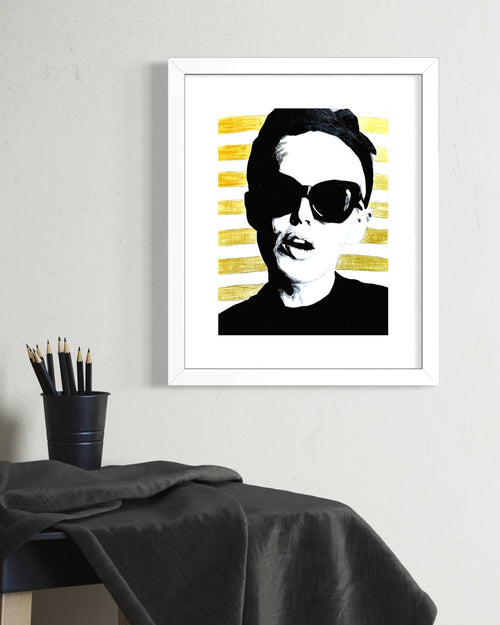 Framed black and white limited edition print with a woman wearing sunglasses and gold stripes, on a wall above a table.