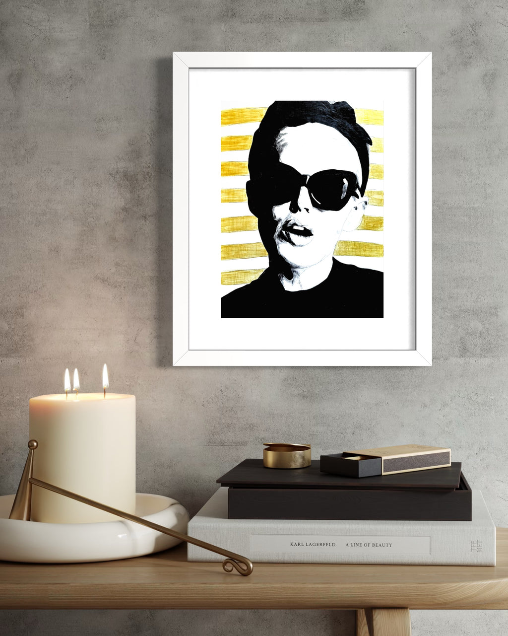 Framed limited edition print of a woman with sunglasses and gold stripes,  on a wall above a table with books and a candle.