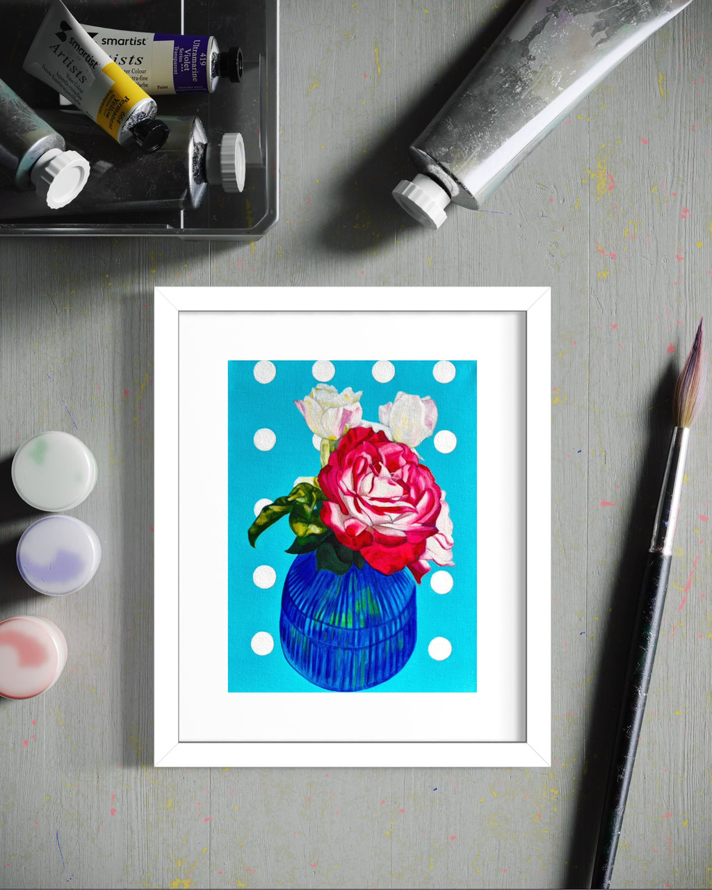 Framed limited edition print of a colourful flower in a blue vase on a grey surface with art supplies.