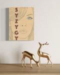 Painting on board with a stylised woman's face and 'SYZYGY' text above two gold deer figurines on a wooden surface.