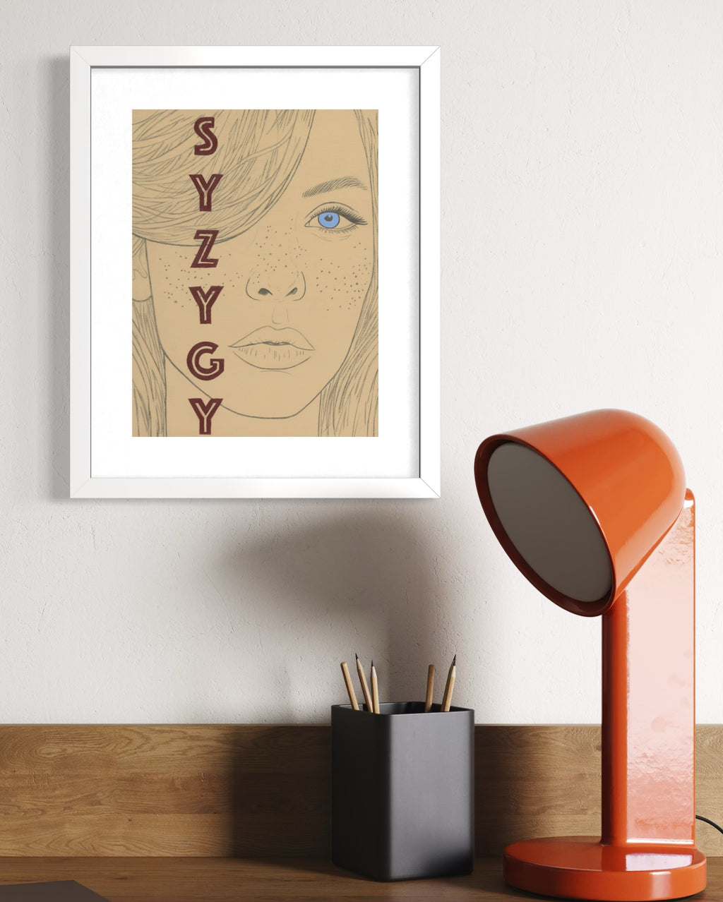 Framed artwork with 'SYZYGY' text and a portrait, next to an orange lamp on a wooden surface.