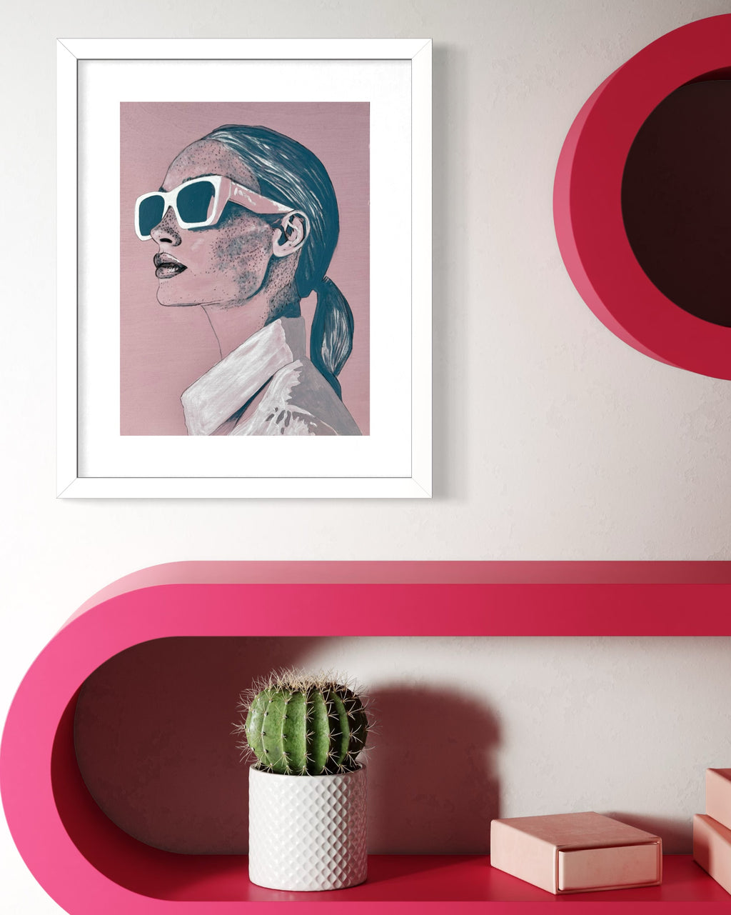 Framed artwork of a woman, 'Frankie', with sunglasses on a pink wall above a pink shelf with a cactus and books.