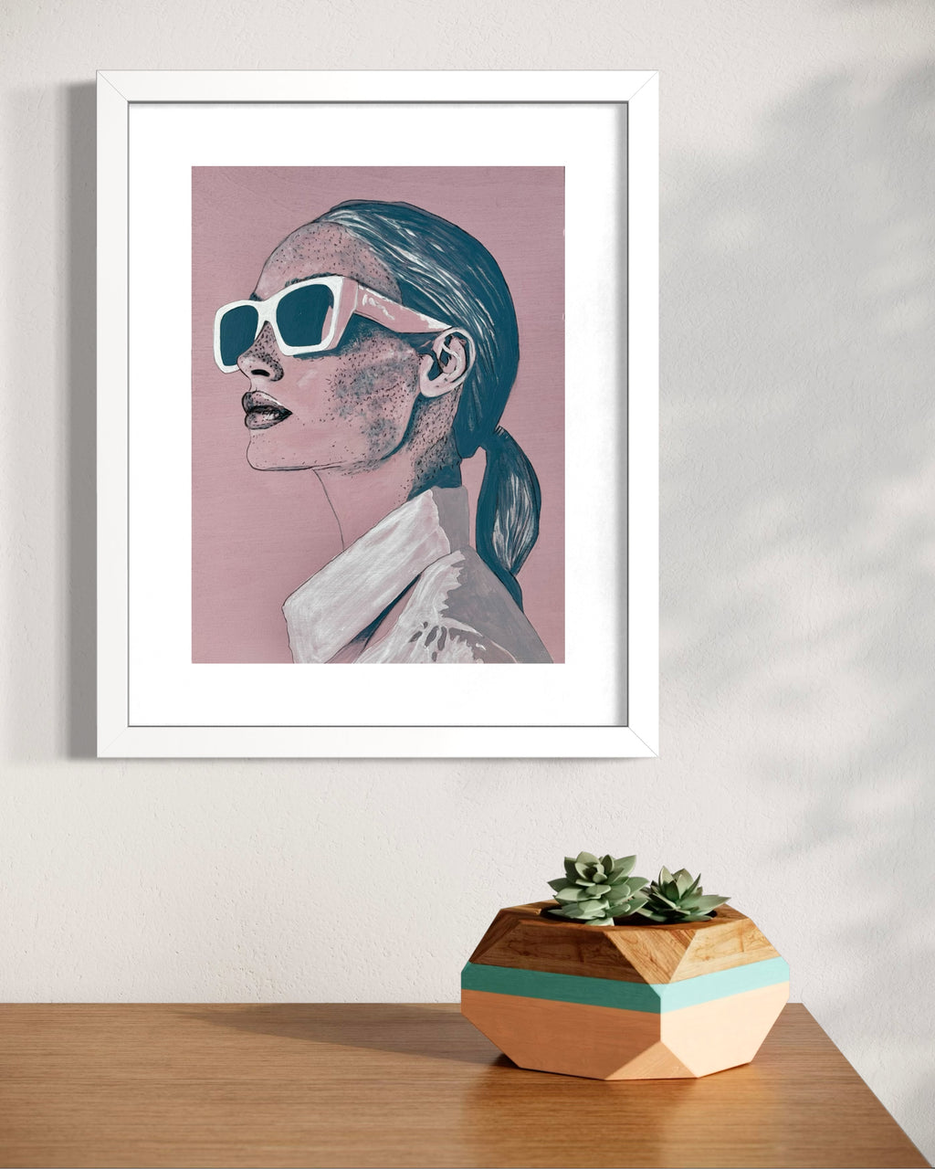 Framed artwork of 'Frankie', a woman wearing sunglasses on a pink background, placed on a wooden surface with a geometric plant pot.