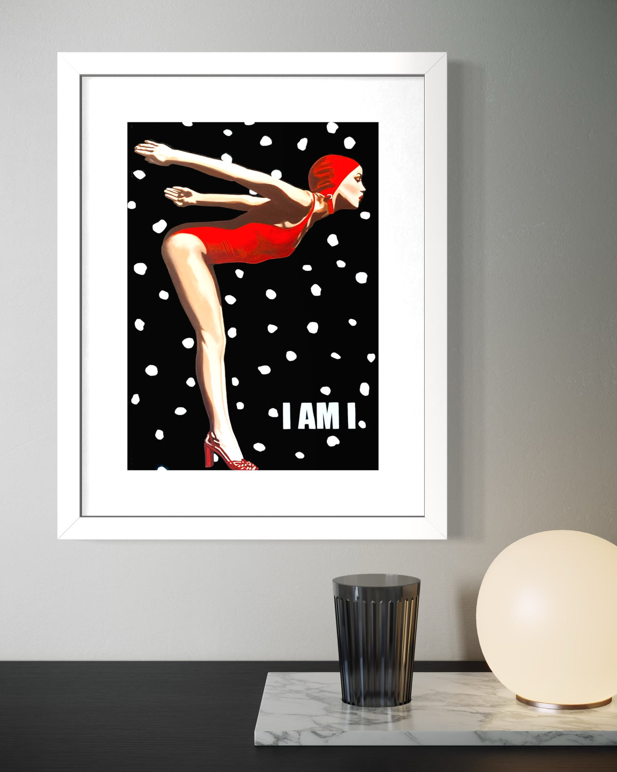 A framed art print featuring a stylized woman in a red swimsuit on a black background with white polka dots and the text 'I AM I.' The white-framed print is displayed on a gray wall above a table with a black cup and a round white lamp.