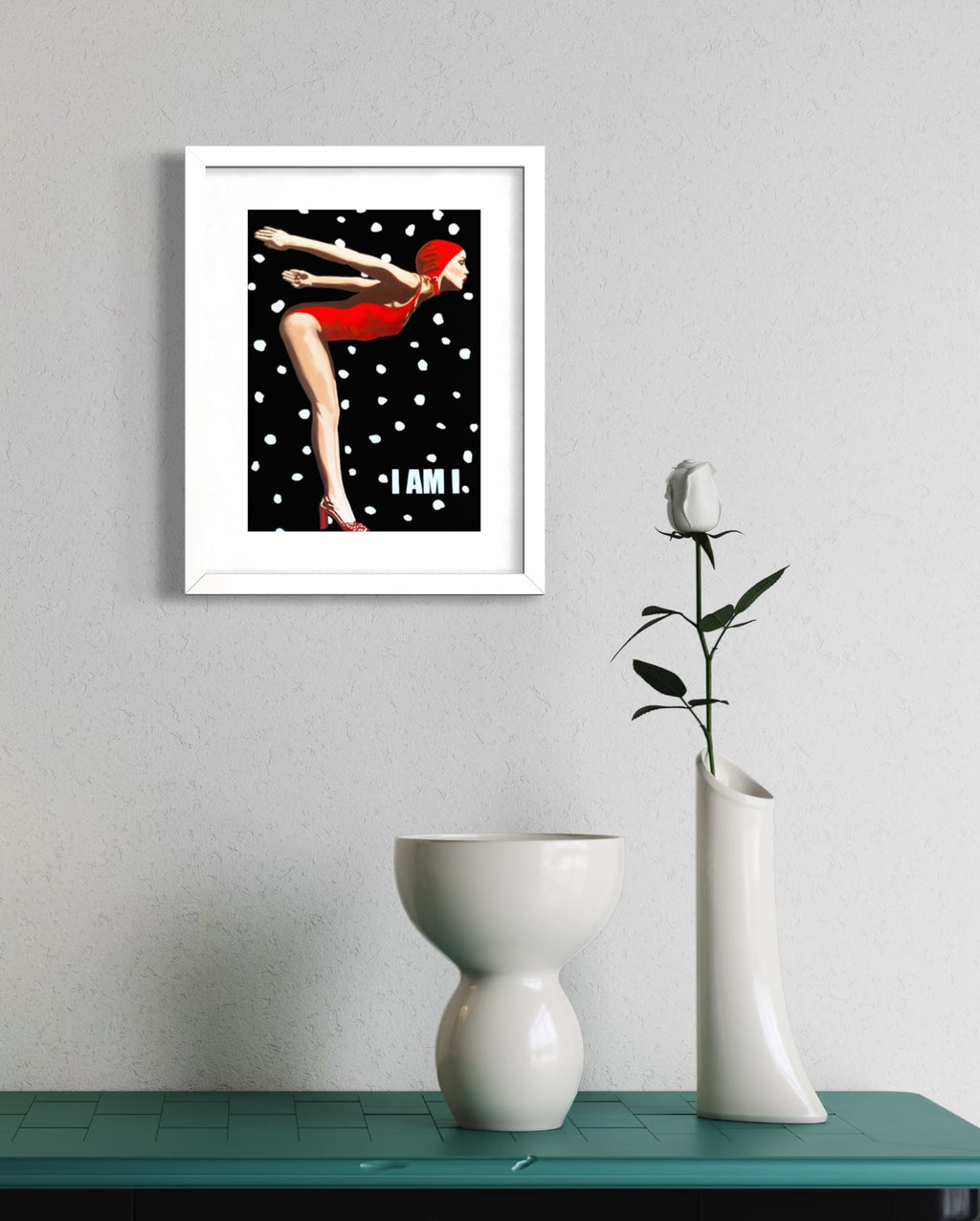 A framed art print featuring a retro-style illustration of a woman in a red swimsuit on a black background with white polka dots and the text 'I AM.', displayed on a wall above a teal table with white ceramic vases.