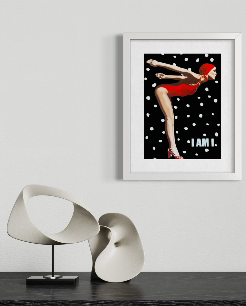 A framed art print featuring a retro illustration of a woman in a red dress against a black background with white polka dots and the text 'I AM I.' The print is displayed on a light gray wall above a dark table with two abstract white sculptures.