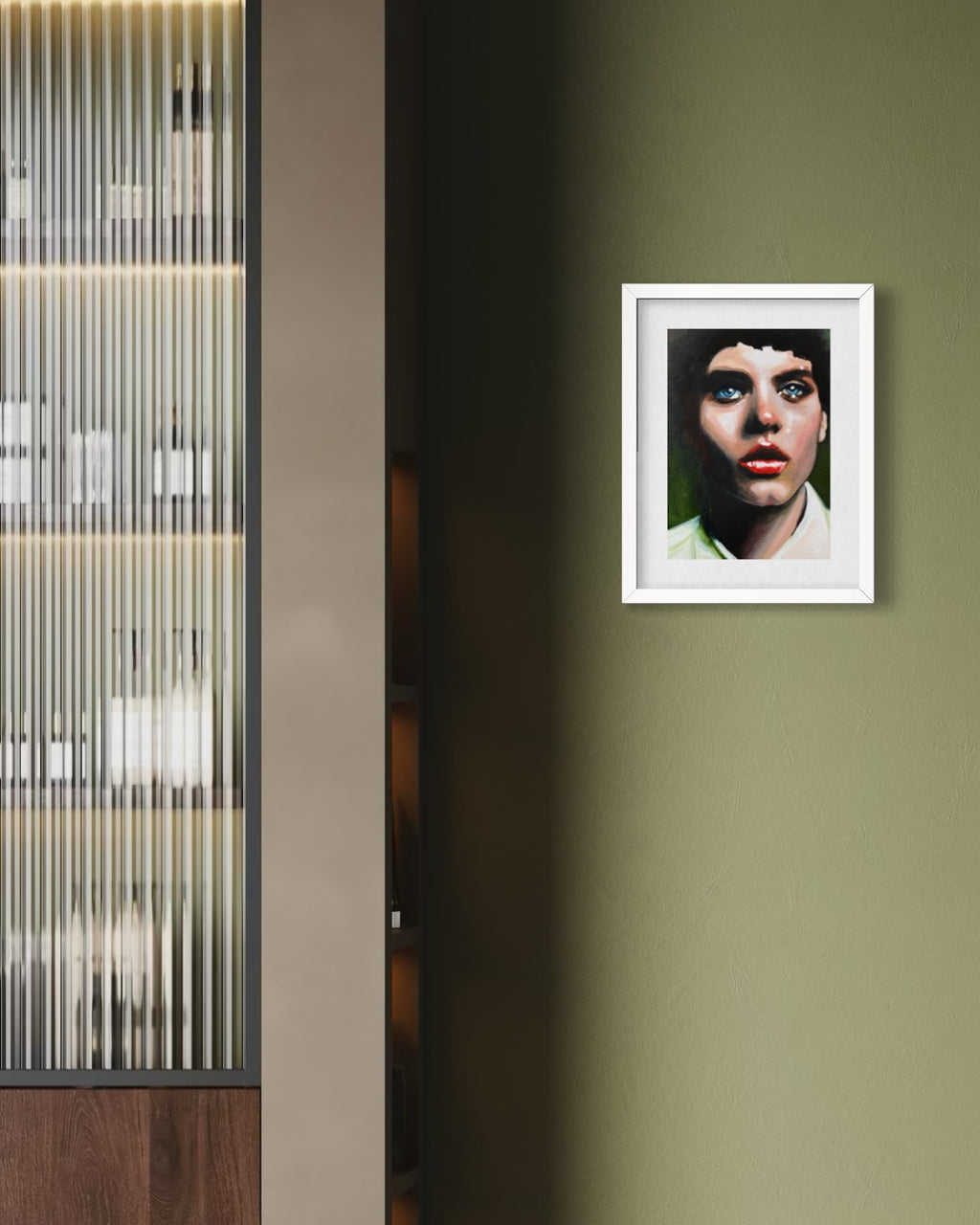 A framed portrait print of a woman with blue eyes hangs on an olive green wall in a modern interior setting with a ribbed glass cabinet on the left.