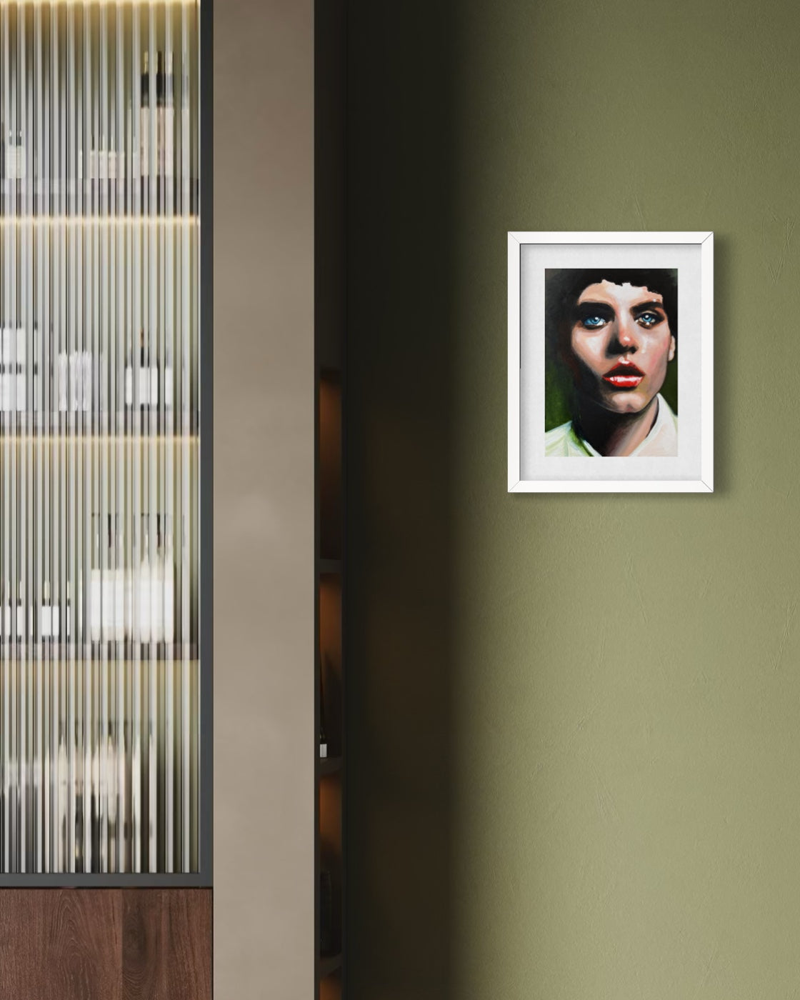 A framed portrait print of a woman with blue eyes hangs on an olive green wall in a modern interior setting with a ribbed glass cabinet on the left.