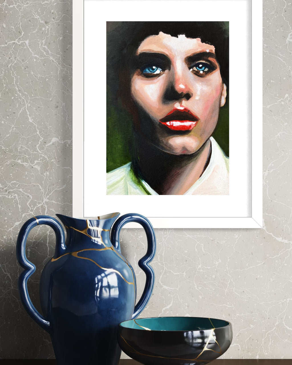 A framed art print featuring a stylized portrait of a woman with blue eyes and red lips, displayed on a textured gray wall above a dark blue ceramic vase and a black bowl.