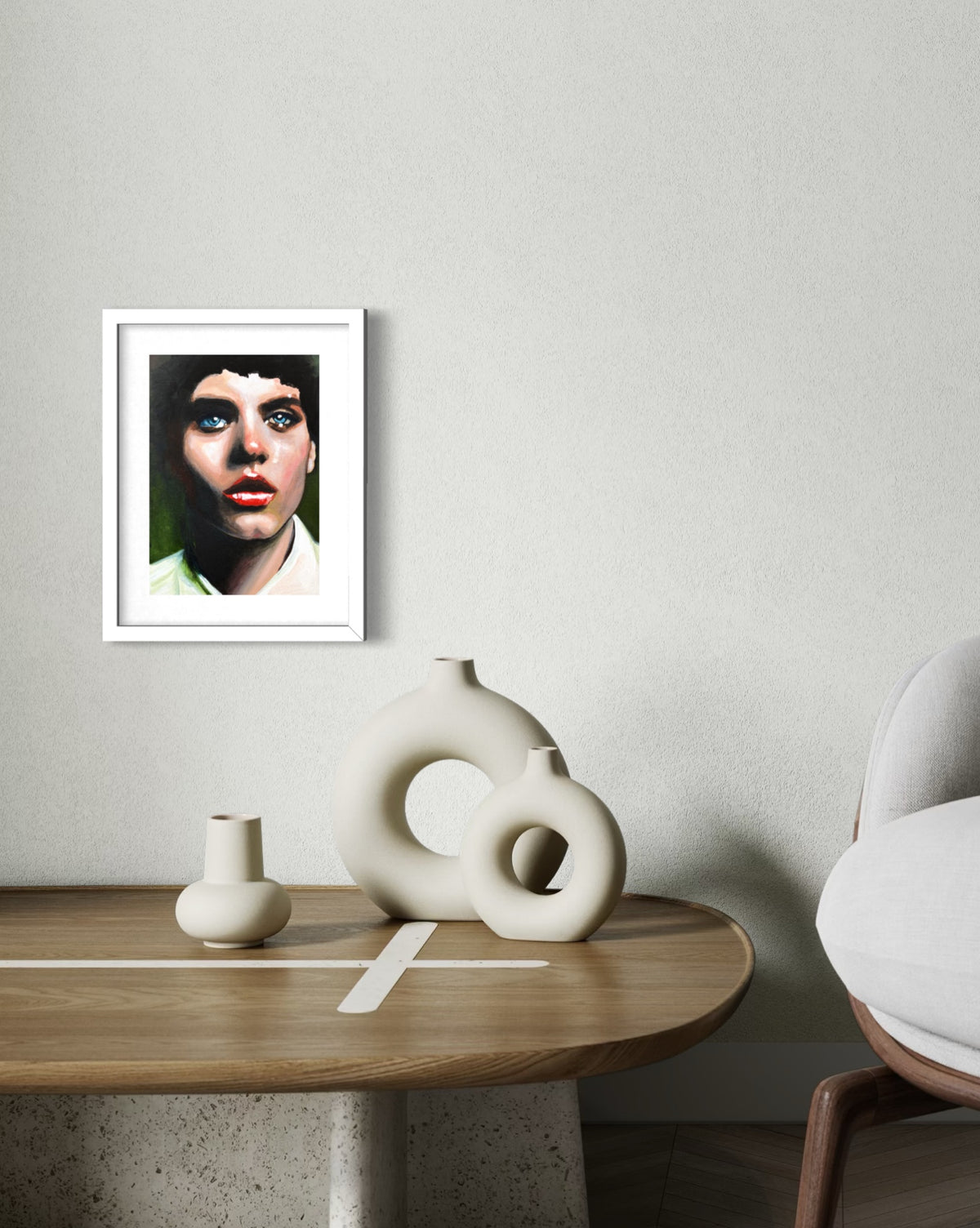 A framed art print of a woman's portrait with dramatic blue eye makeup, displayed on a light gray wall above a wooden table with modern ceramic vases.