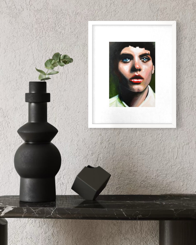 A framed art print featuring a stylized portrait of a woman with blue eyes and red lips, displayed on a textured light gray wall above a black marble console table with a black vase and decorative object.
