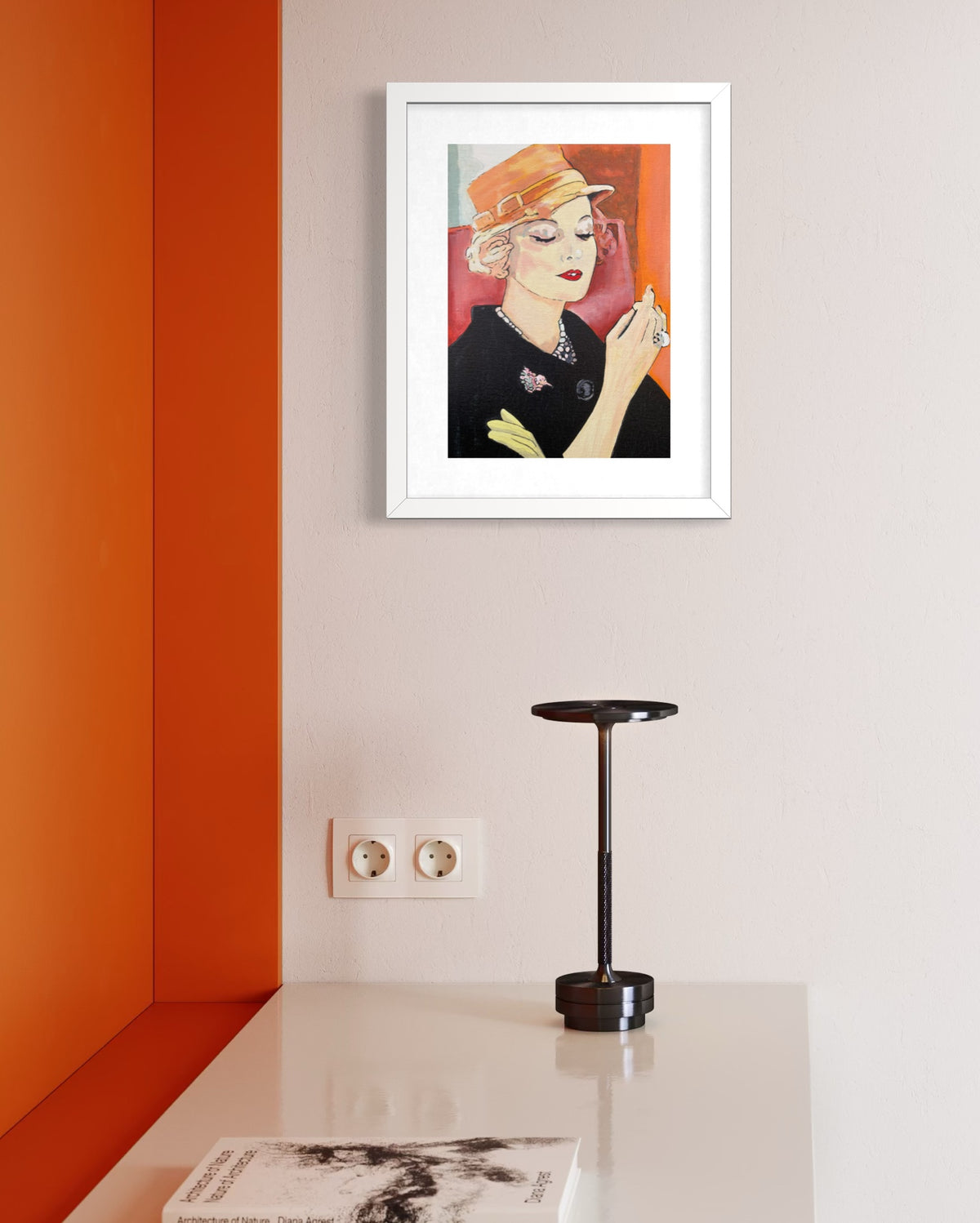 A framed art print featuring a stylized illustration of a woman in a black outfit and orange hat, displayed on a light gray wall above a white table with a black lamp and a book.