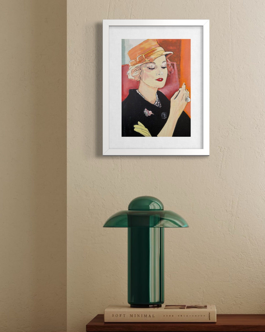 A framed art print featuring a stylized portrait of a woman in a black outfit and orange hat, displayed on a beige wall above a green table lamp on a wooden surface.
