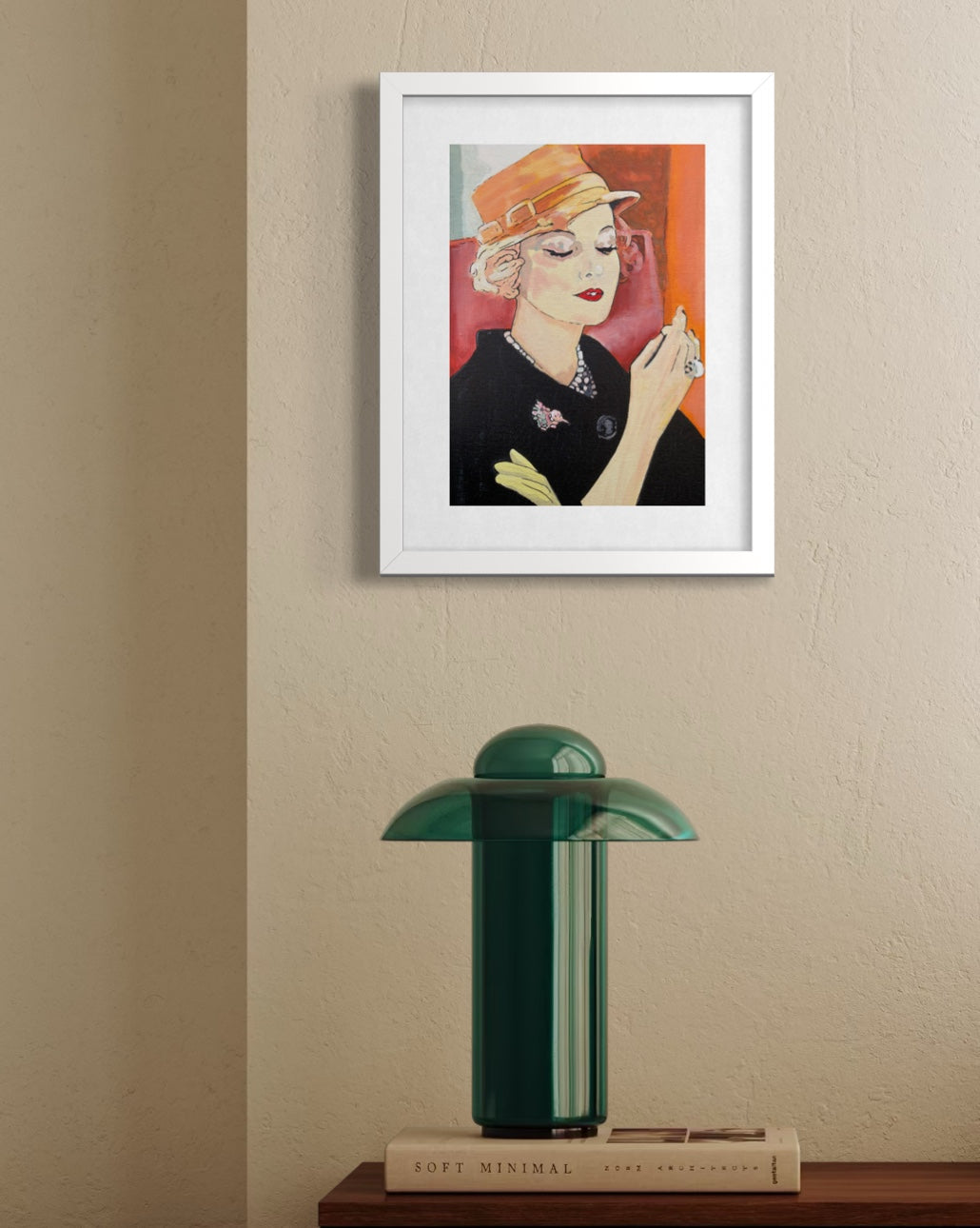 A framed art print featuring a stylized portrait of a woman in a black outfit and orange hat, displayed on a beige wall above a green table lamp on a wooden surface.