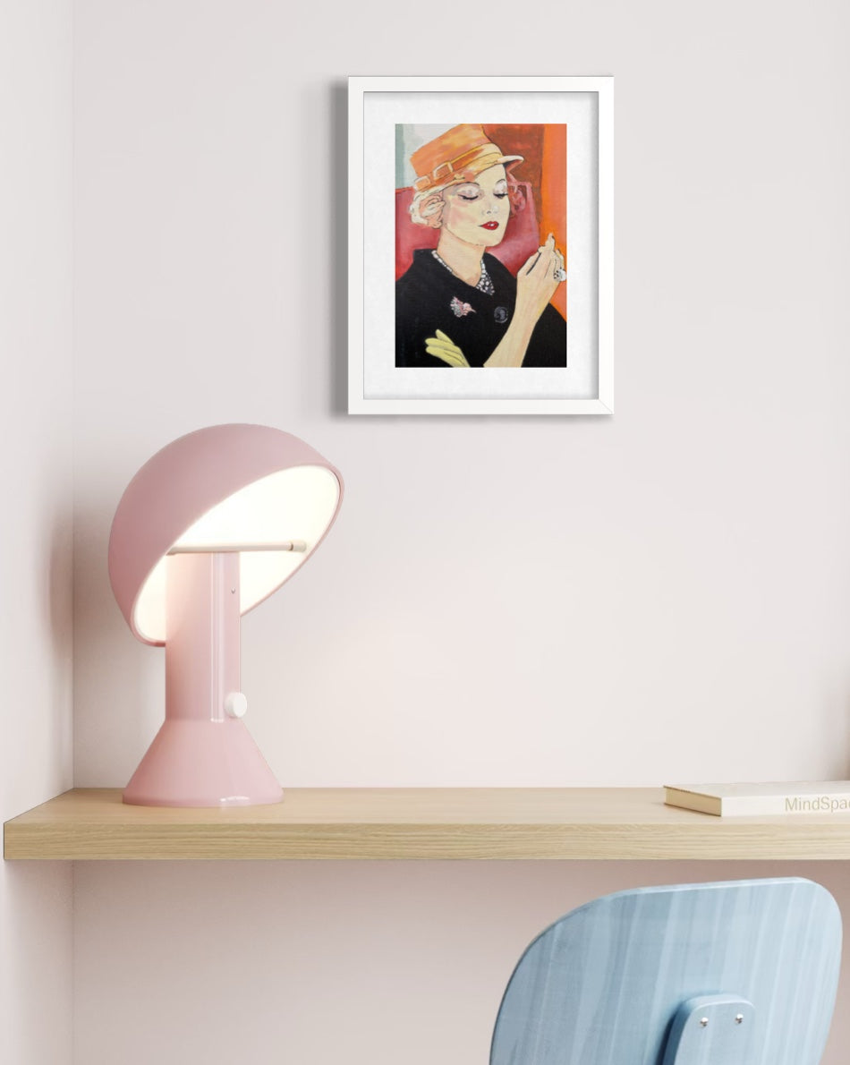 A framed art print featuring a stylized portrait of a woman in a black outfit and orange hat, hanging on a light pink wall above a wooden desk with a pink lamp and a blue chair.