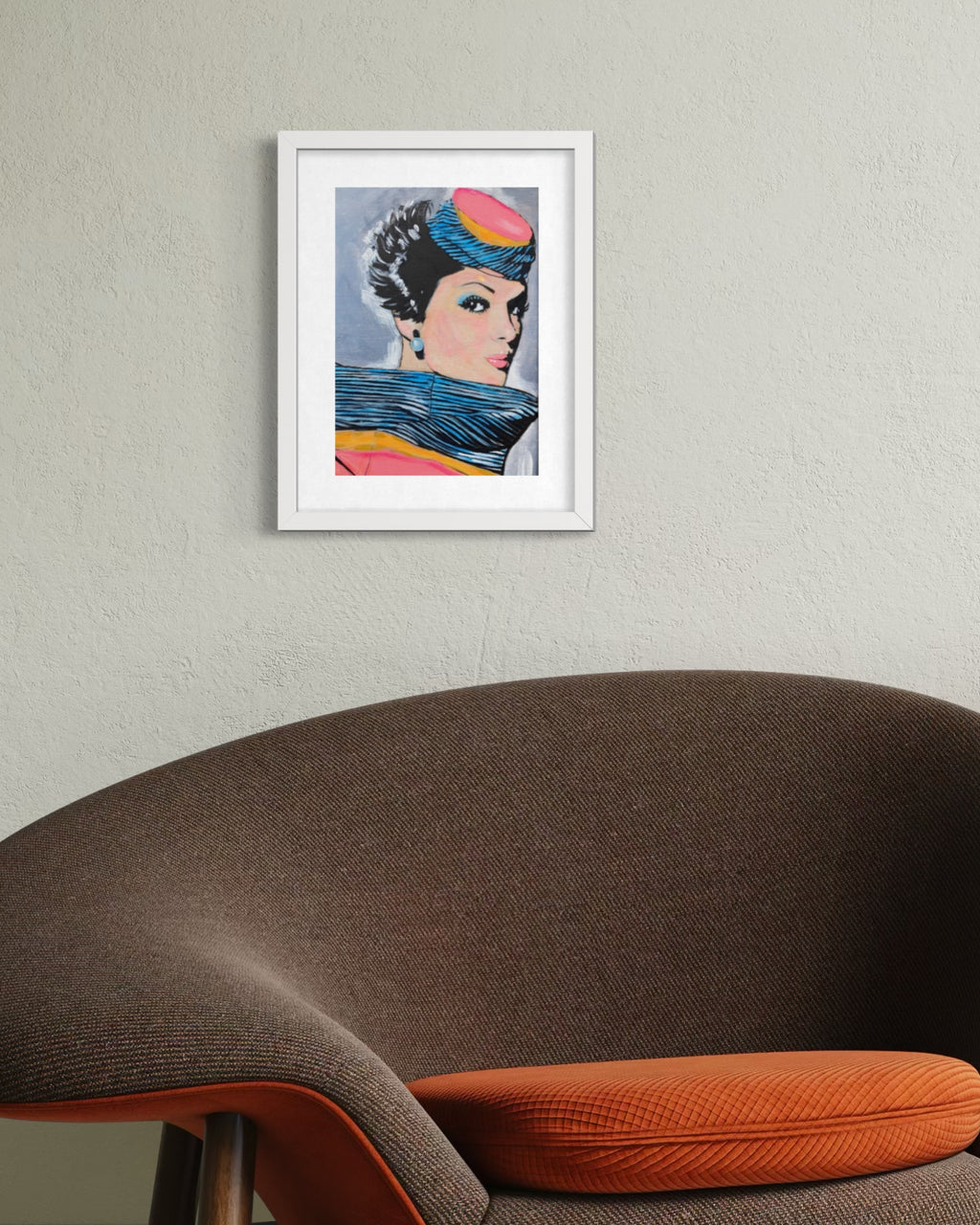 A framed art print featuring a stylized pop-art portrait of a woman with a colorful headpiece and blue scarf, displayed on a textured light gray wall above a brown upholstered chair with an orange cushion.