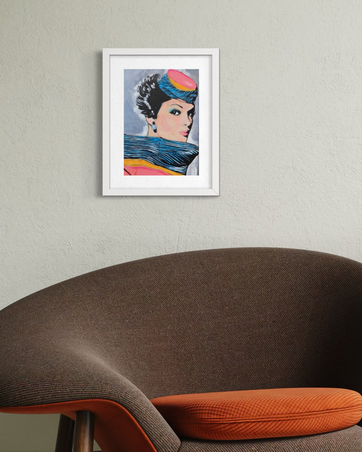 A framed art print featuring a stylized pop-art portrait of a woman with a colorful headpiece and blue scarf, displayed on a textured light gray wall above a brown upholstered chair with an orange cushion.
