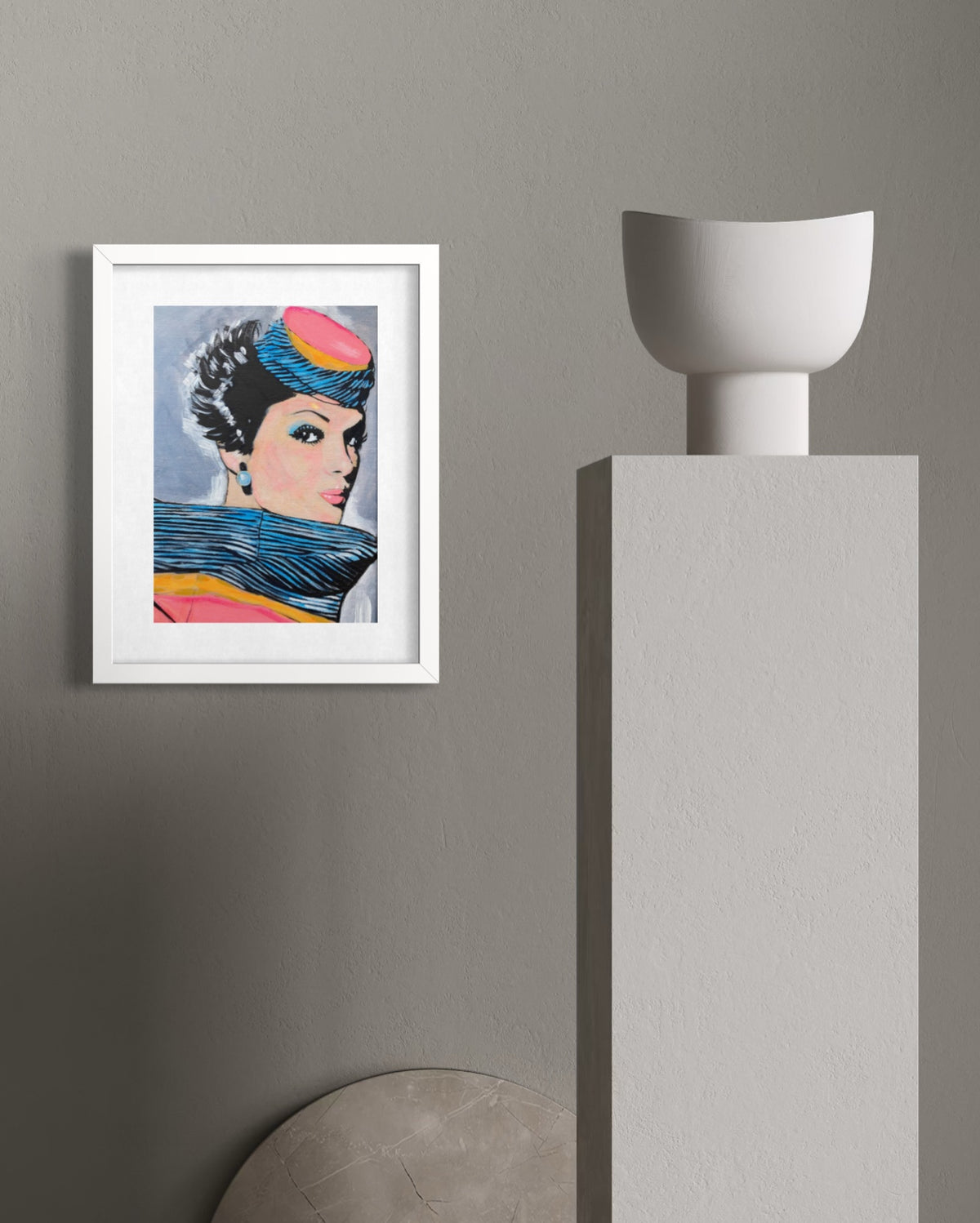 A framed art print featuring a colorful pop-art portrait of a woman, displayed on a textured gray wall next to a modern white pedestal.
