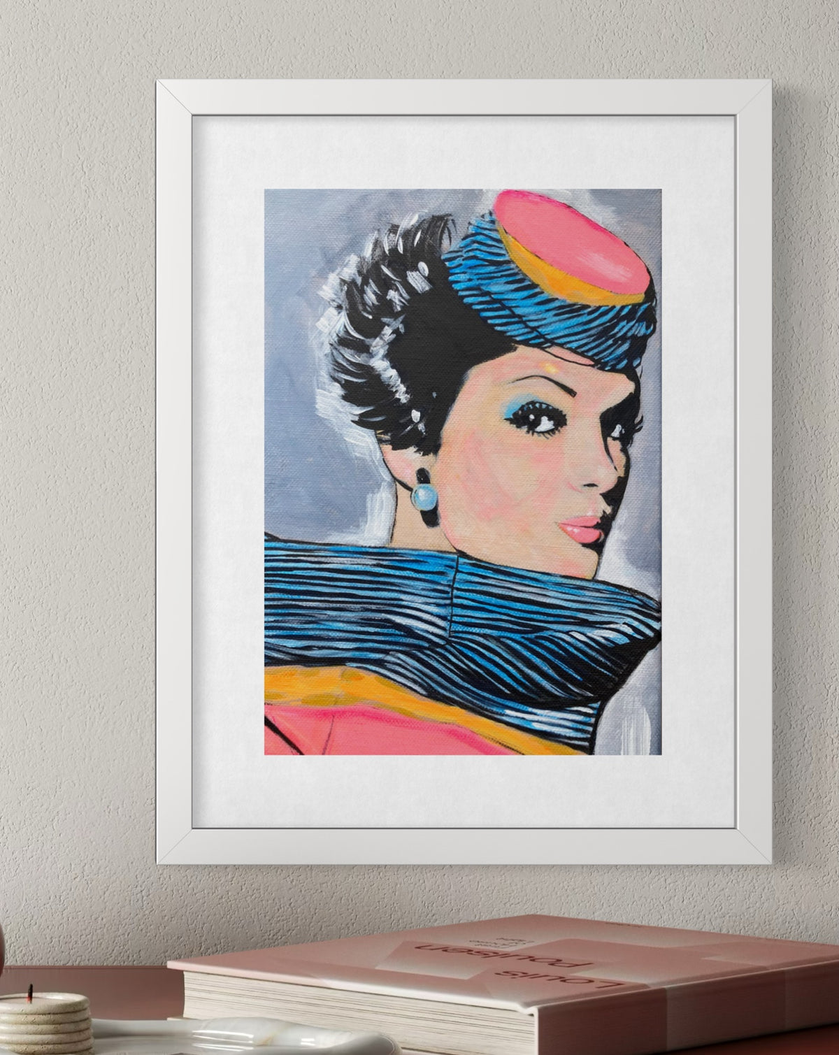 A framed pop-art portrait print of a woman with bold black, blue, pink, and yellow details, displayed on a wall above a book.