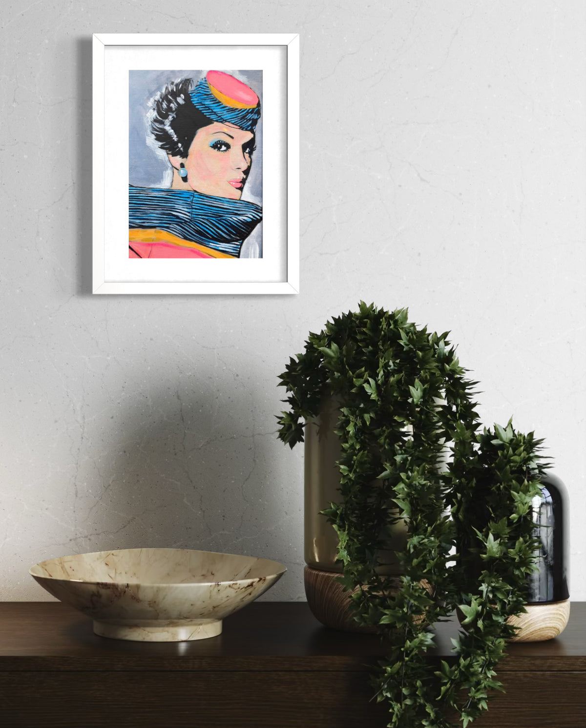 A framed pop-art print of a woman with a colorful headwrap hanging on a light gray textured wall above a dark wooden console table. The print is in a white frame, displayed next to a ceramic bowl and a potted trailing green plant.