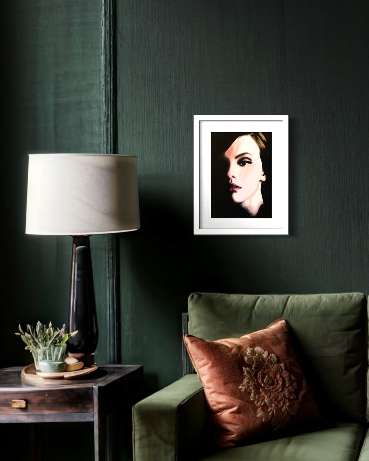 A framed black and white portrait print of a woman's face hangs on a dark green textured wall above an olive green armchair with a brown embroidered pillow. A black table lamp with a beige shade sits on a dark wooden side table next to the chair.