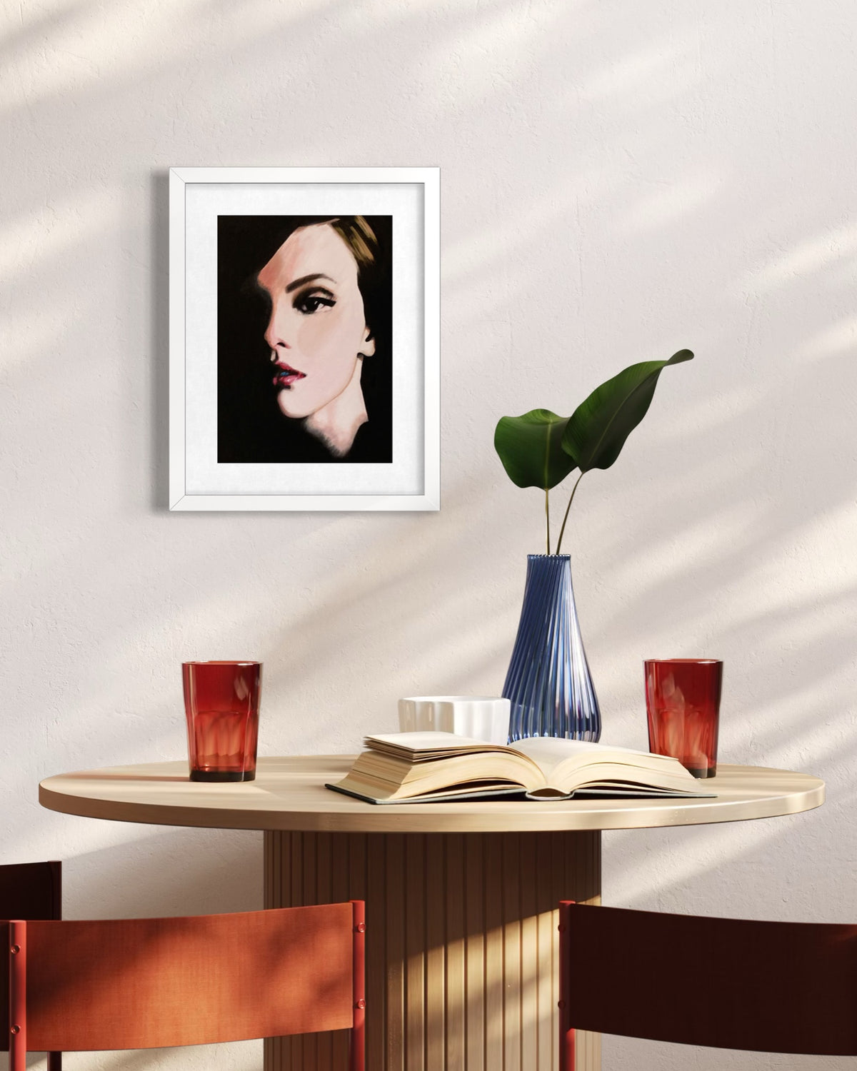 A framed art print featuring a stylized portrait of a woman in black and white, hung on a light beige wall above a wooden dining table with chairs, an open book, and a blue vase with a green leaf.