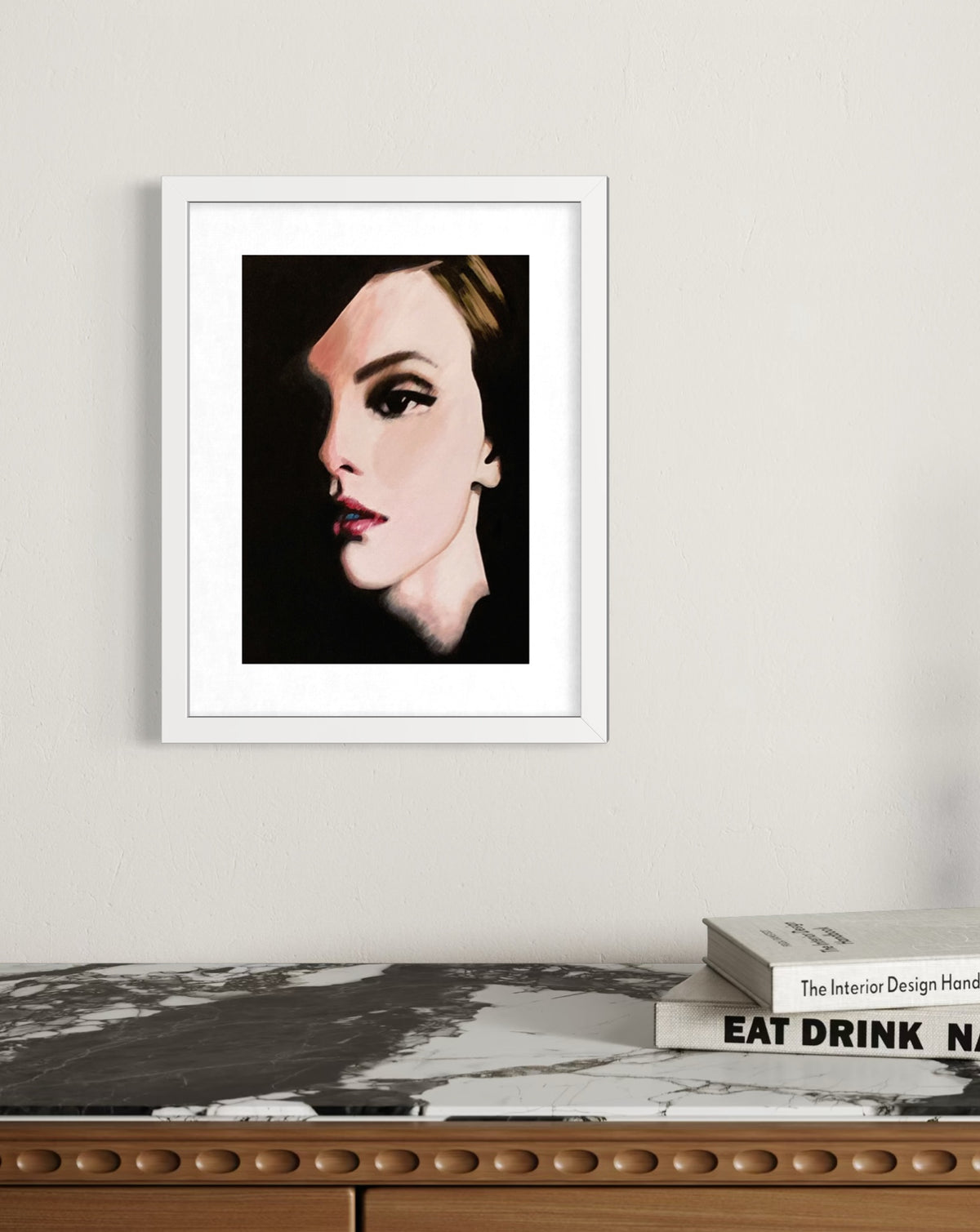 A framed art print featuring a stylized portrait of a woman in black and beige tones, displayed on a light wall above a marble-topped console table with stacked books.