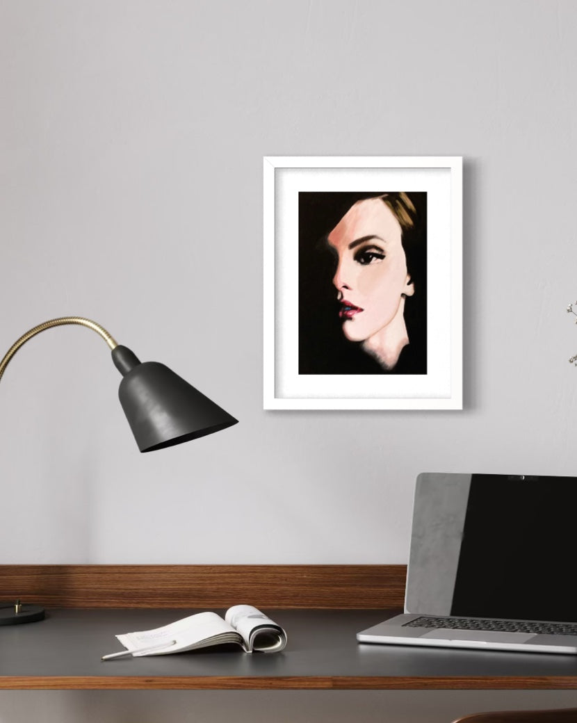 A framed art print featuring a stylized portrait of a woman with dark hair and red lips, set against a black background and enclosed in a white frame. The frame is displayed on a light gray wall above a desk with a laptop, an open notebook, and a black desk lamp.