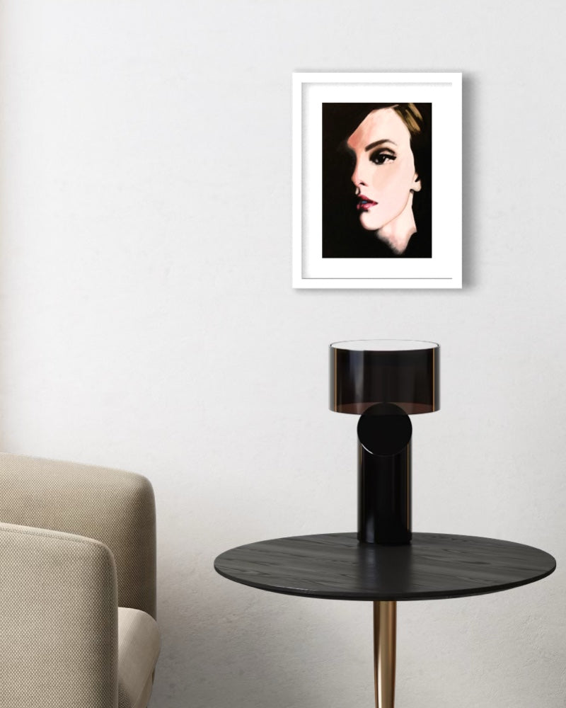 A framed art print featuring a stylized portrait of a woman with dramatic eye makeup, displayed on a light gray wall above a black side table with a modern lamp.