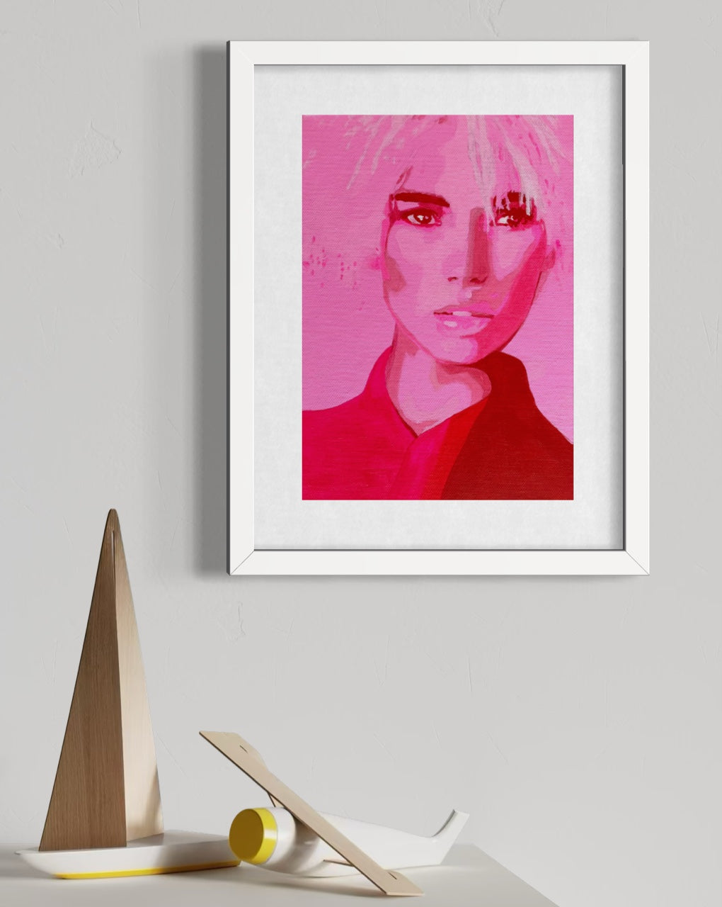 A framed art print featuring a stylized portrait of a woman in vibrant pink and red tones, displayed on a light gray wall above a small decorative model ship.