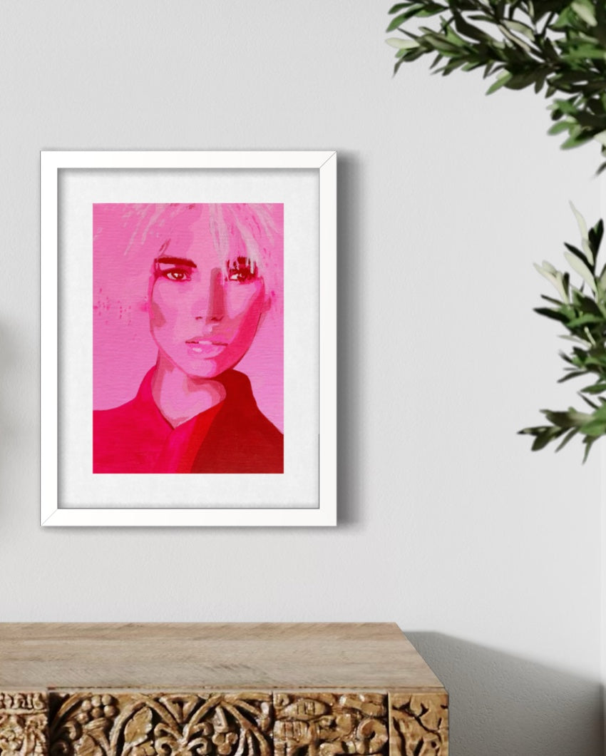 A framed art print featuring a stylized portrait of a woman in vibrant pink and red tones, displayed on a light gray wall above a carved wooden console table with green foliage in the corner.
