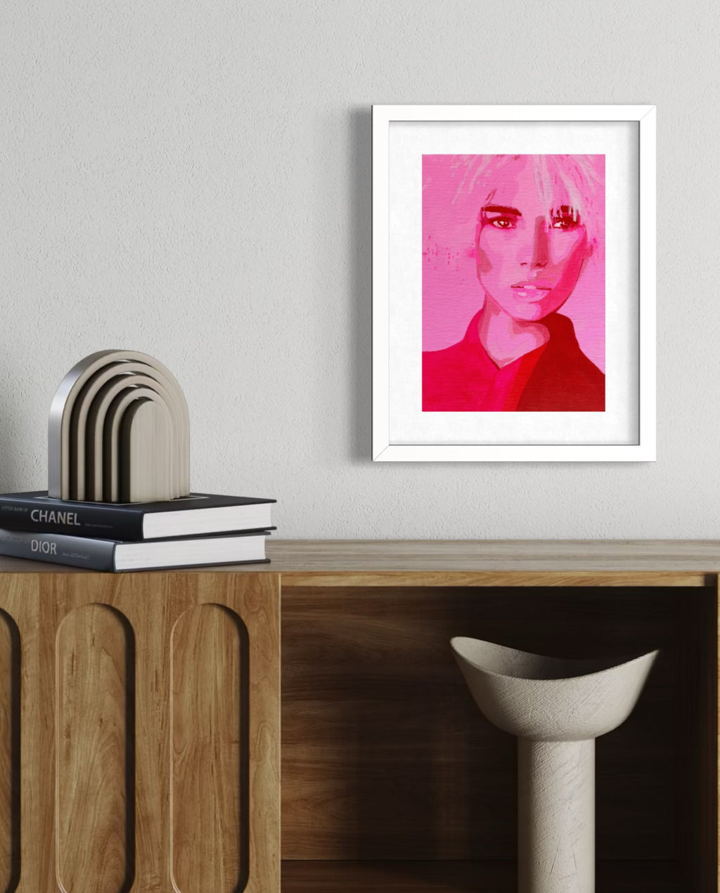 A framed pop-art portrait print of a woman in vibrant pink and red tones, displayed on a light gray wall above a wooden console table with books and a stool.