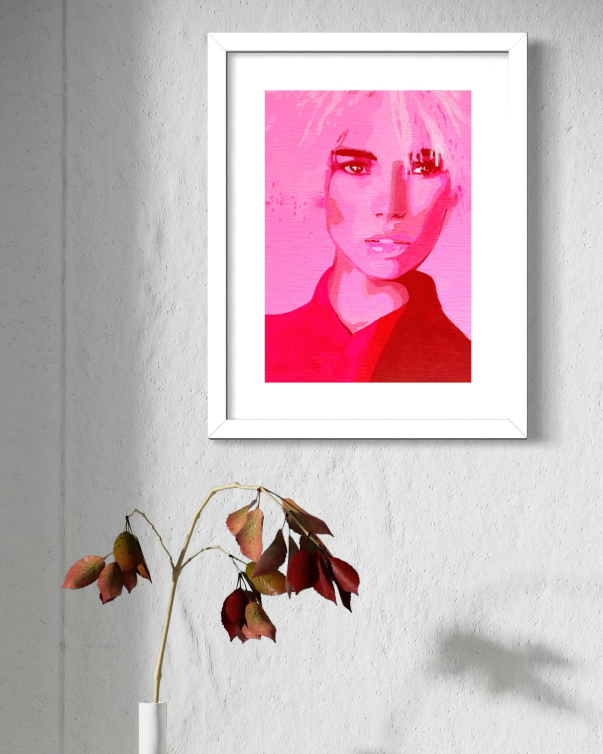 A framed art print featuring a stylized portrait of a woman in vibrant pink and red tones, displayed on a textured light gray wall above a minimalist vase with dried leaves.