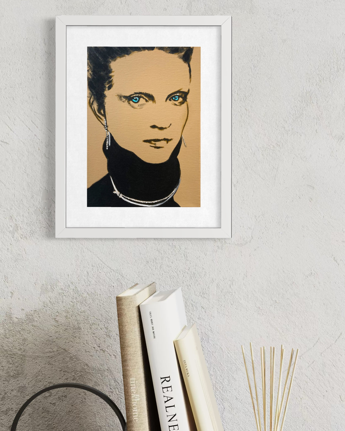 A framed pop-art style portrait print of a woman with blue eyes, shown hanging on a textured light gray wall above stacked books and a reed diffuser.