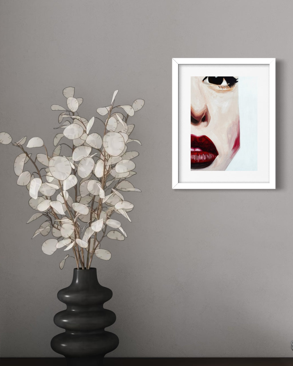 A framed art print featuring a close-up portrait of a woman with red lips and black sunglasses, displayed on a gray wall next to a black vase with dried white eucalyptus branches.