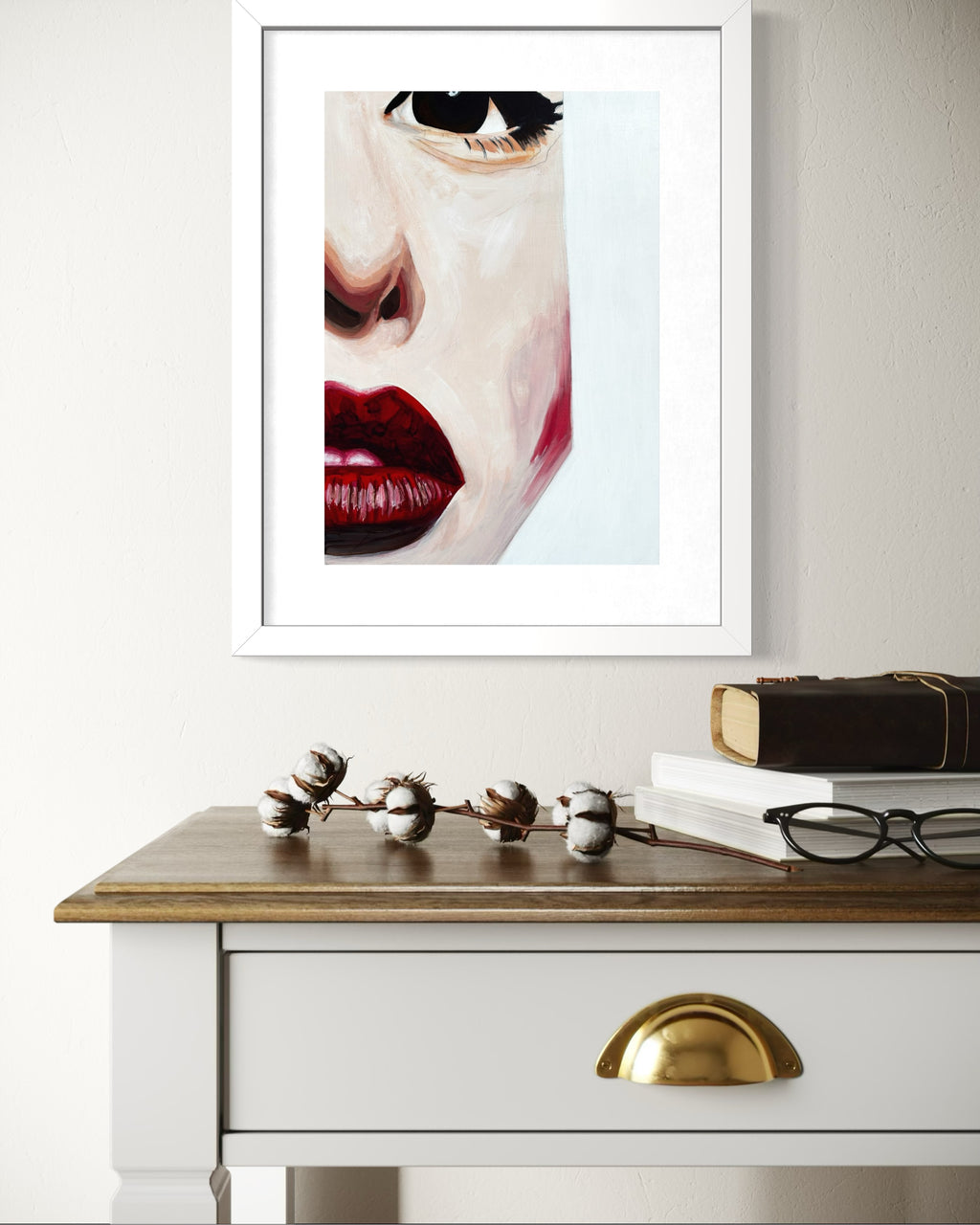 A framed art print featuring a close-up portrait of a woman's face with bold red lips, displayed on a white wall above a wooden console table with books and glasses.