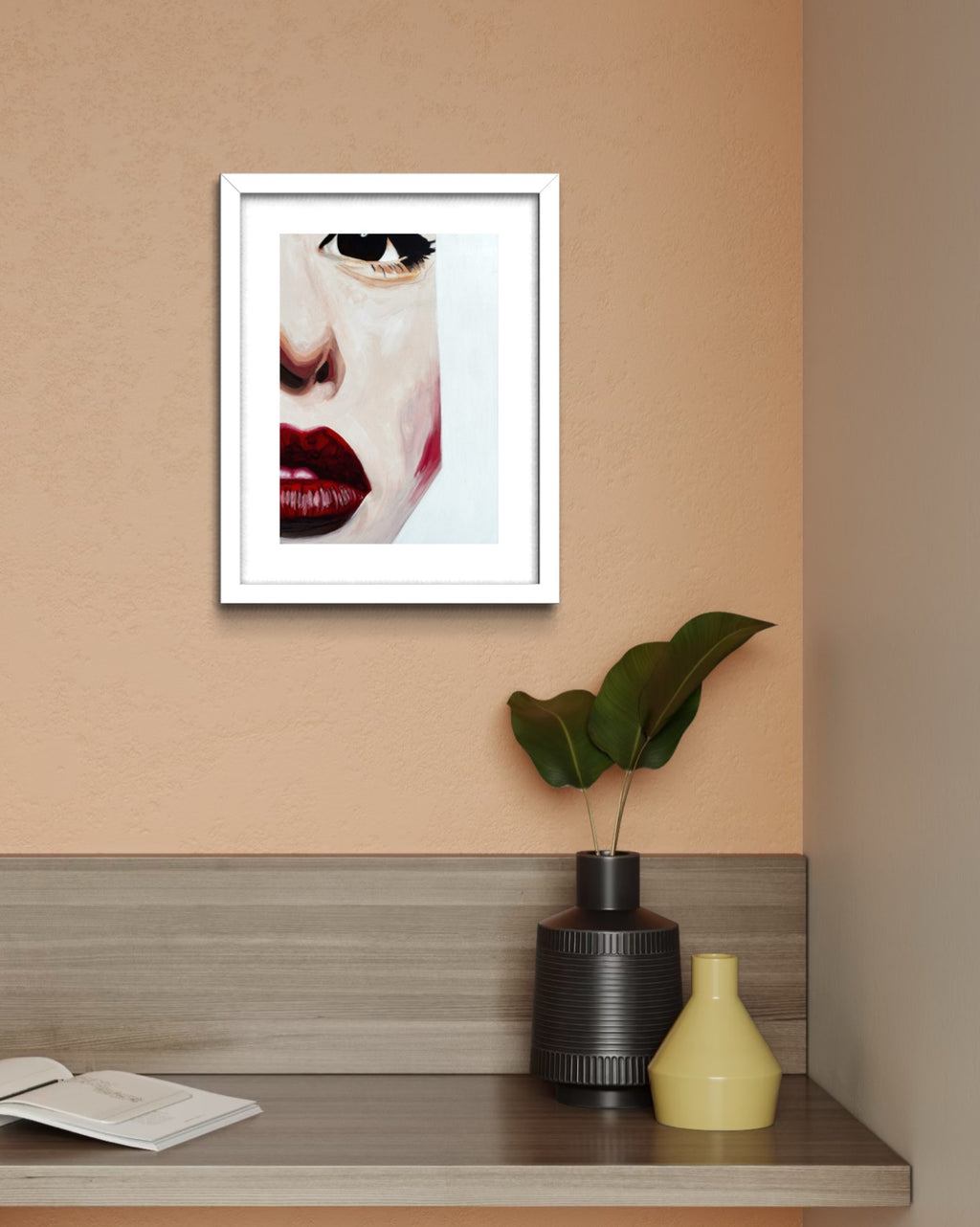 A framed art print featuring a close-up portrait of a woman's face with red lips and black sunglasses, displayed on a peach-colored wall above a wooden shelf with decorative vases and an open book.