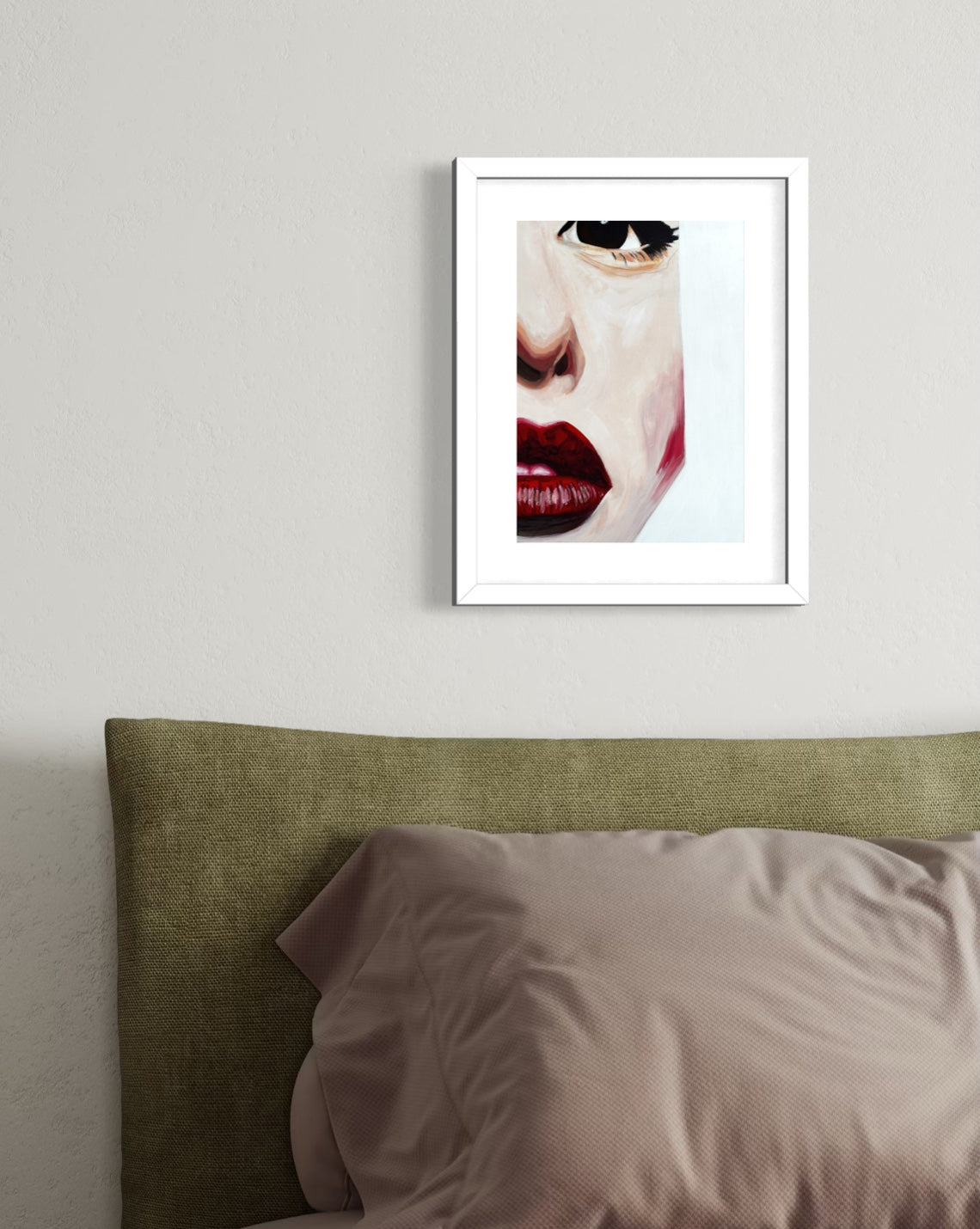 A framed art print featuring a close-up portrait of a woman's face with red lips and dark eye makeup, displayed on a light gray wall above a bed with an olive green headboard and mauve pillows.