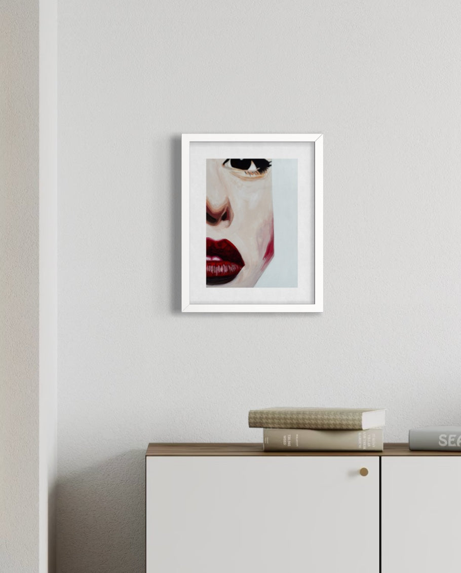 A framed art print featuring a close-up portrait of a woman's face with red lips and dark sunglasses, displayed on a light gray wall above a white cabinet with books.