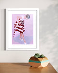 A framed art print featuring a stylized illustration of a woman in a red and white striped dress, hanging on a light beige wall above a wooden table with a geometric succulent pot.