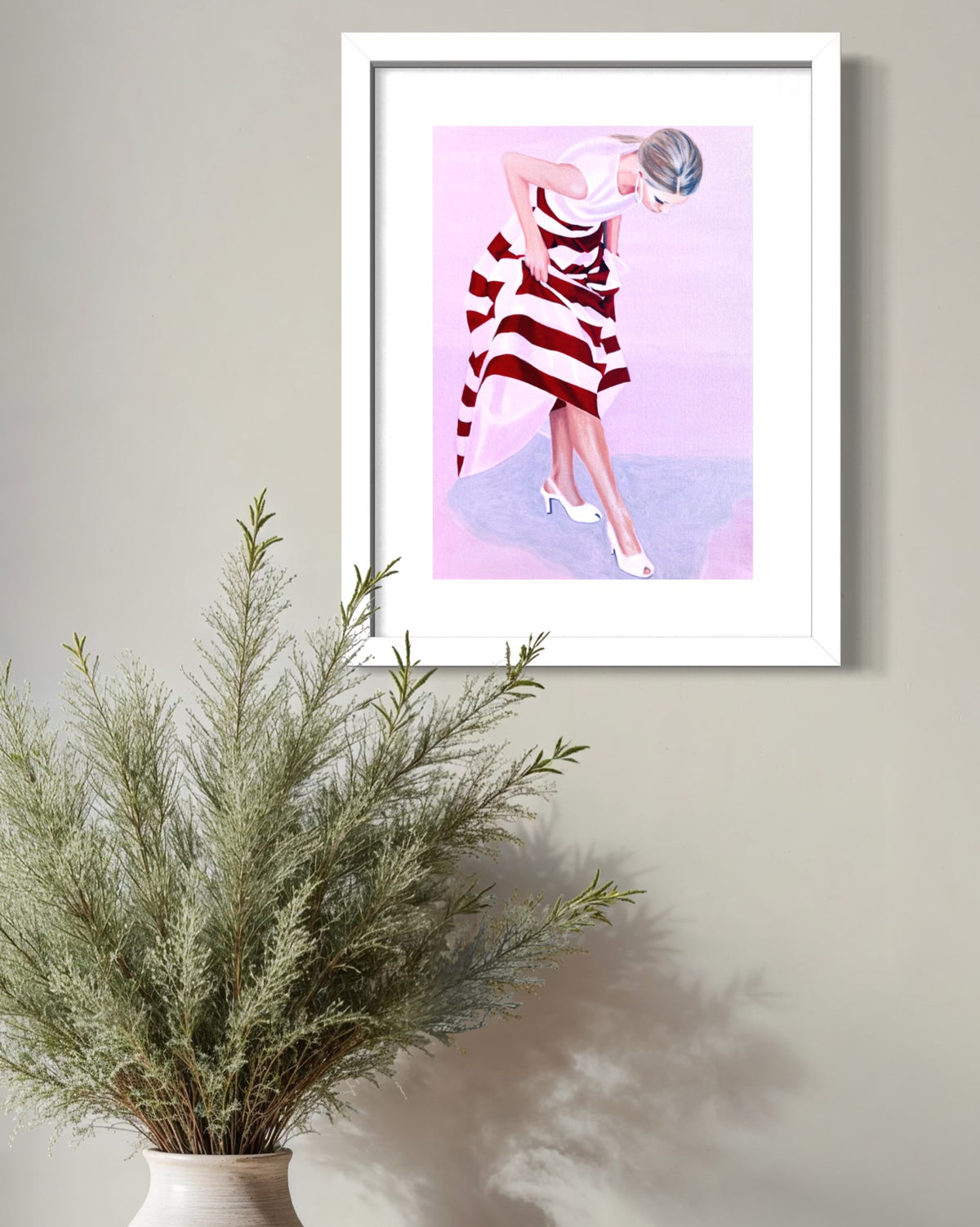 A framed art print featuring a stylized illustration of a woman in a red and white striped dress, hanging on a light gray wall above a potted green plant.