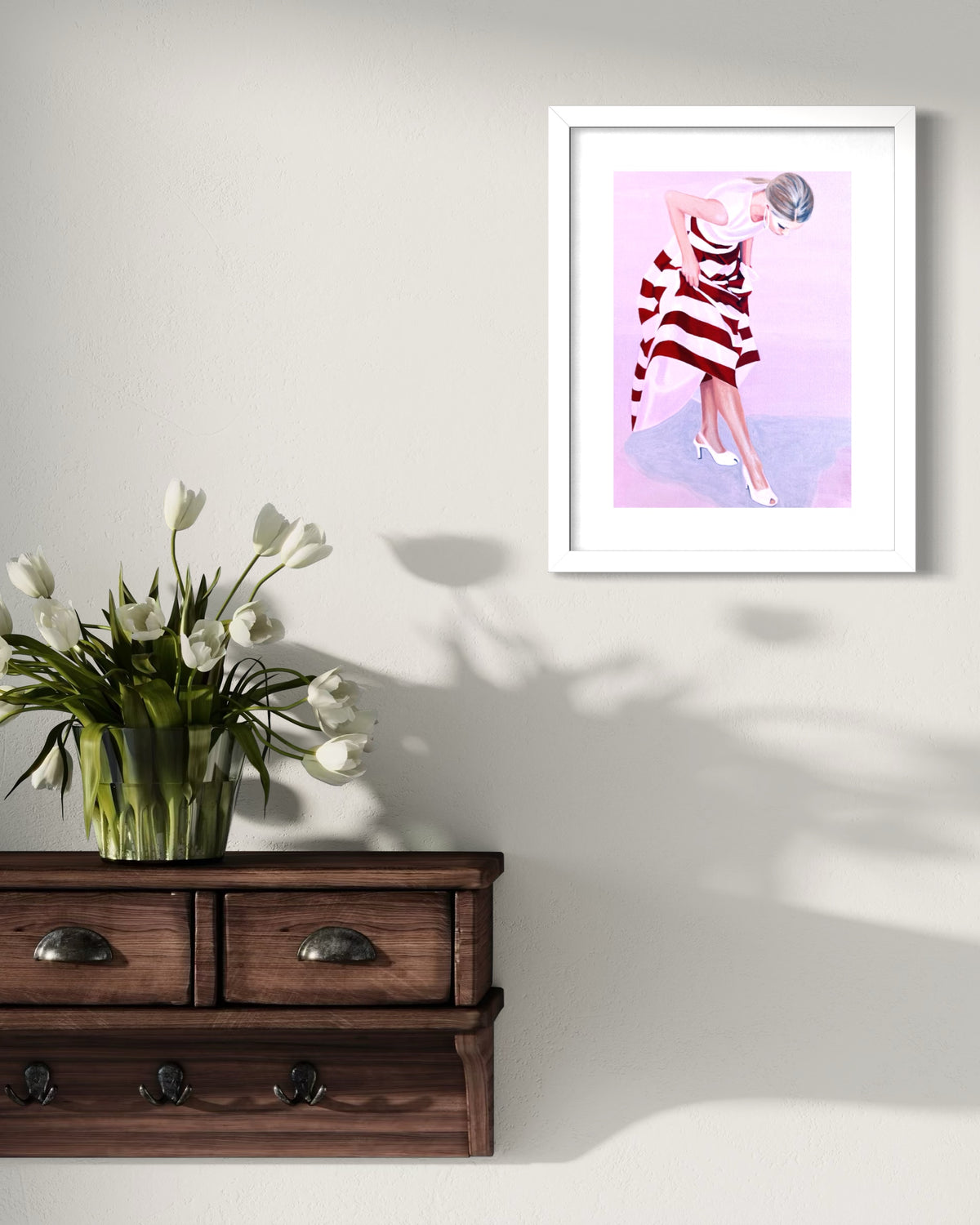 A framed art print featuring a woman in a red and white striped dress against a pink and purple background, displayed on a light gray wall above a dark wooden console table with a vase of white tulips.