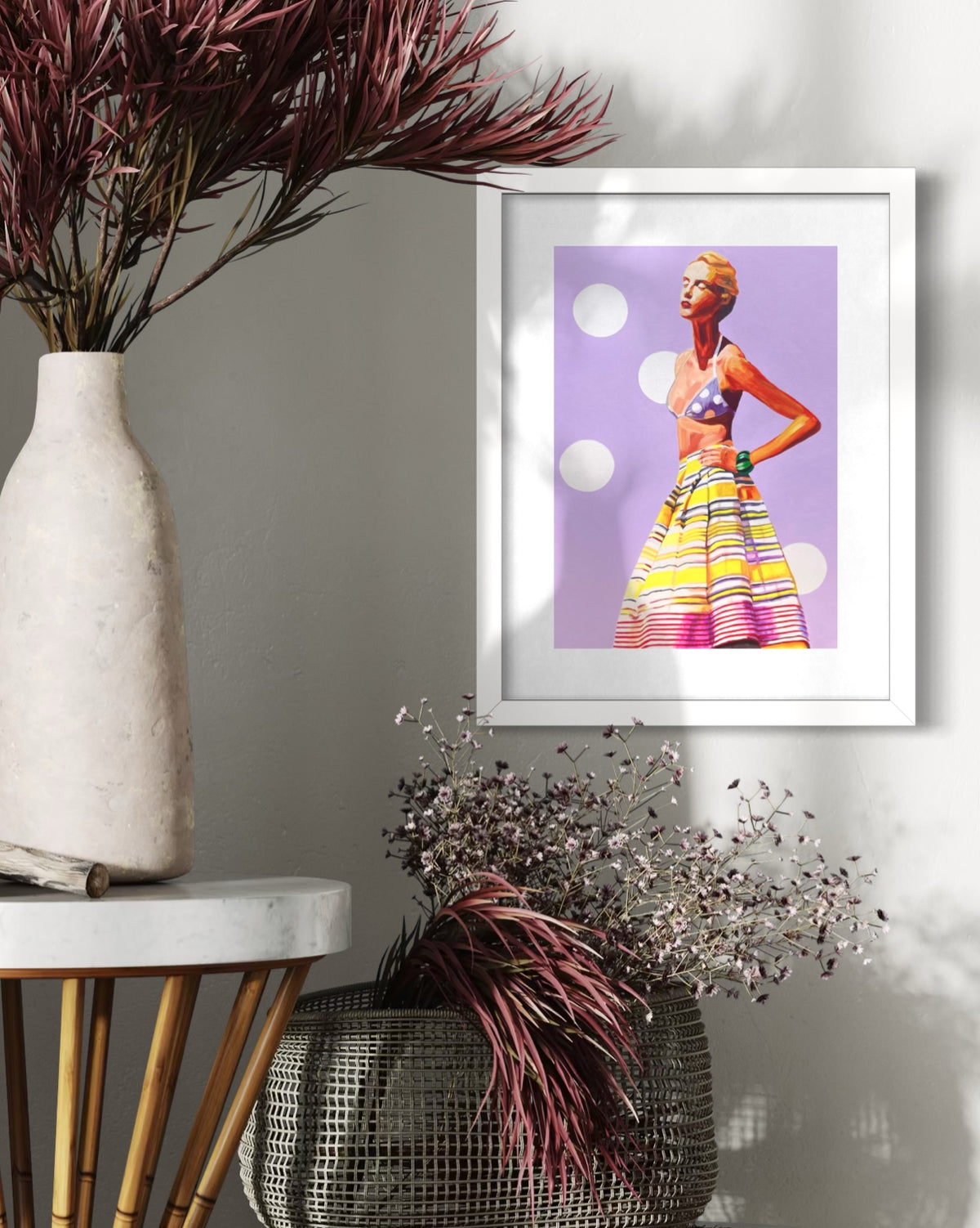 A framed art print featuring a colorful retro-style fashion illustration of a woman against a lavender background, styled on a wall next to a white vase with dried burgundy foliage and a metal basket with dried white flowers.