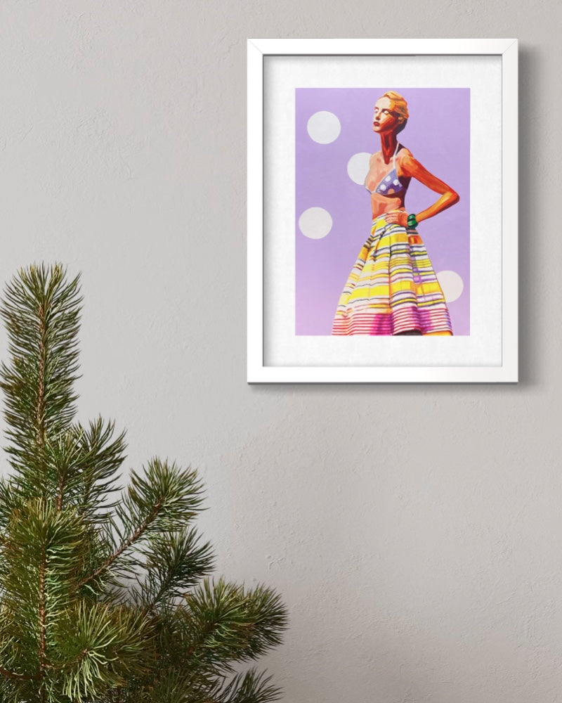 A framed art print featuring a stylized fashion illustration of a woman in a colorful striped skirt against a lavender background, displayed on a light gray wall next to a green pine branch.