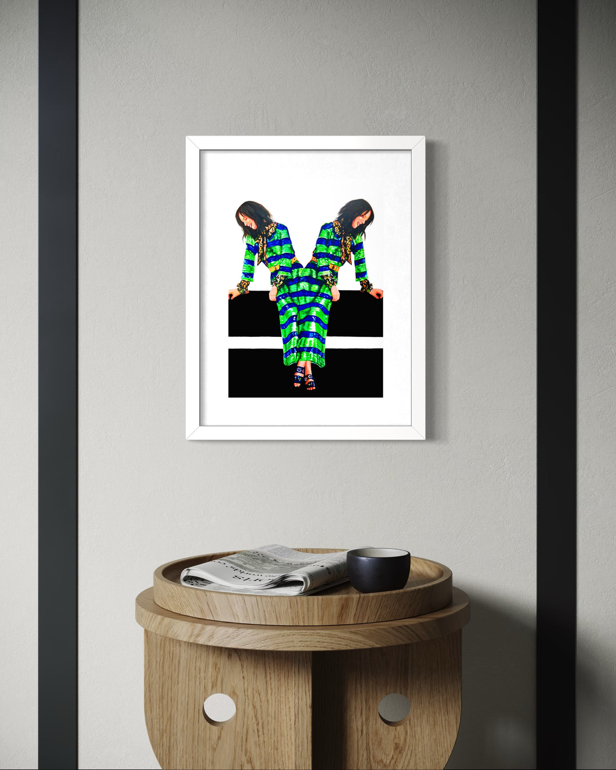 A framed art print featuring a colorful, patterned figure in a green and blue outfit against a black and white background, displayed on a light gray wall above a wooden side table.