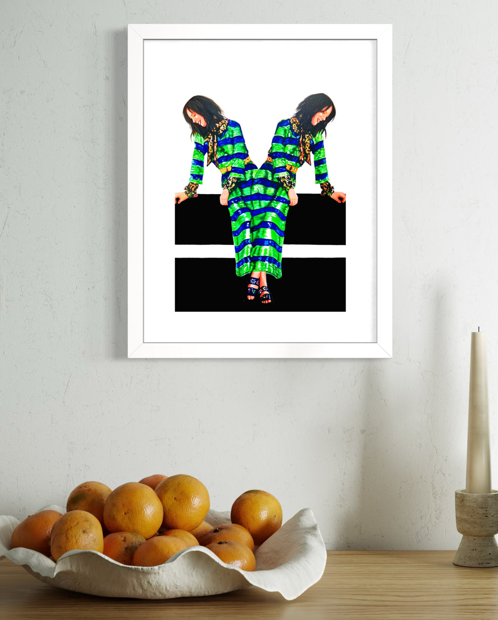 A framed wall art print hangs on a light textured wall above a wooden table. The print shows a mirrored fashion photograph of a woman in a green and black patterned dress.