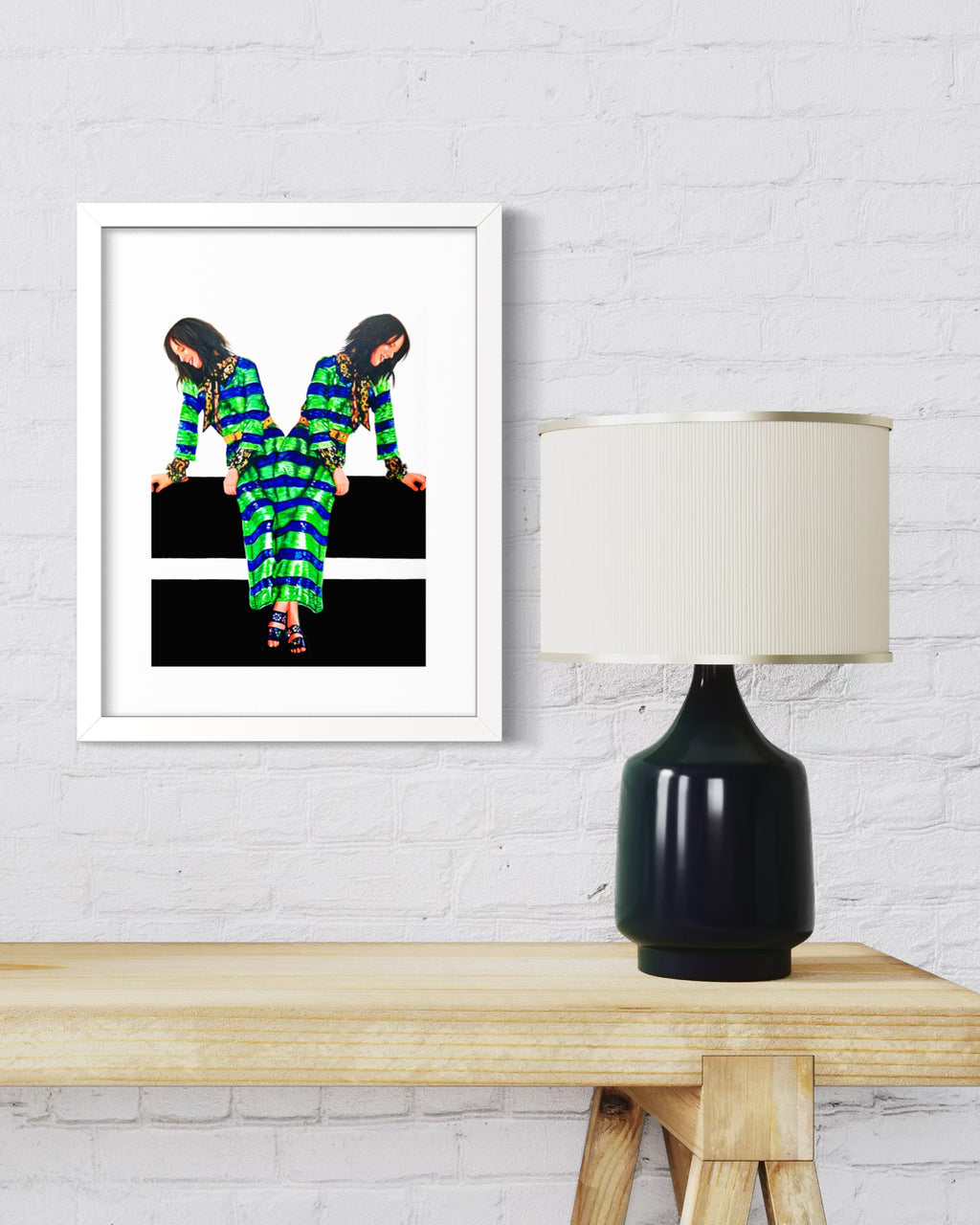 A framed art print featuring a stylized, mirrored fashion illustration of a woman in a green and black patterned outfit, displayed on a white brick wall above a wooden table with a black table lamp.