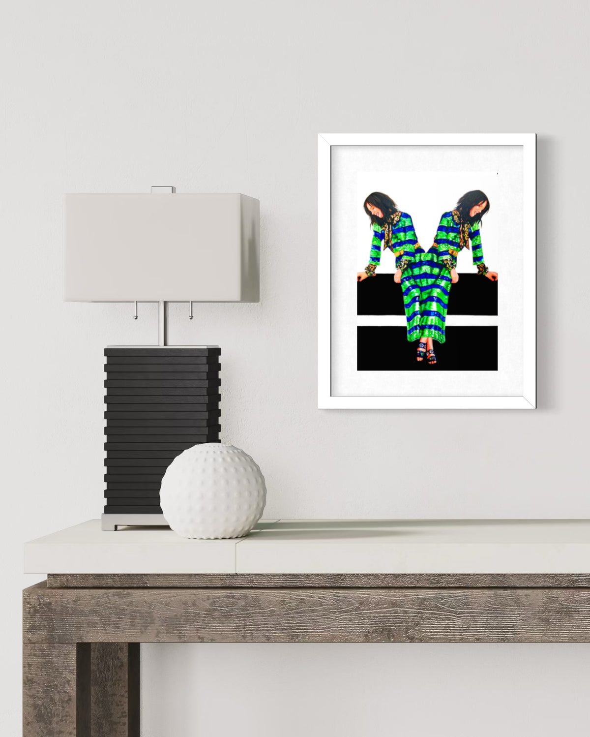 A framed art print featuring a colorful, patterned figure on a black background, displayed on a white wall above a console table with a lamp and a white decorative sphere.